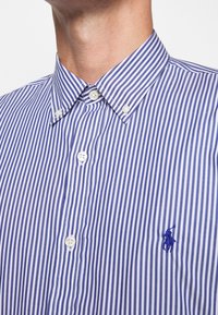 Sininen ja valkoinen pystysuora raitapaita, jossa on kaulus ja tummansininen brodeerattu polo-pelaajalogotaskussa.