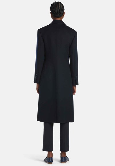 Long manteau bleu marine avec épaules structurées, tissu texturé et une fente au dos, associé à un pantalon assorti et des chaussures plates.