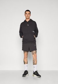 Felpa nera con logo, pantaloni corti abbinati e sneaker nere con dettagli verdi. Calze bianche in vista. Il modello è posizionato davanti a uno sfondo neutro.