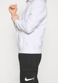 Vit hoodie med framficka, justerbar dragsko och ribbade mudd. Kombinerad med svarta cargobyxor med en Nike-logotyp.