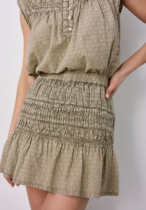 Femme portant une robe sans manches beige texturée avec taille smockée, inserts en dentelle et ourlet à volants, debout avec une main sur la hanche.