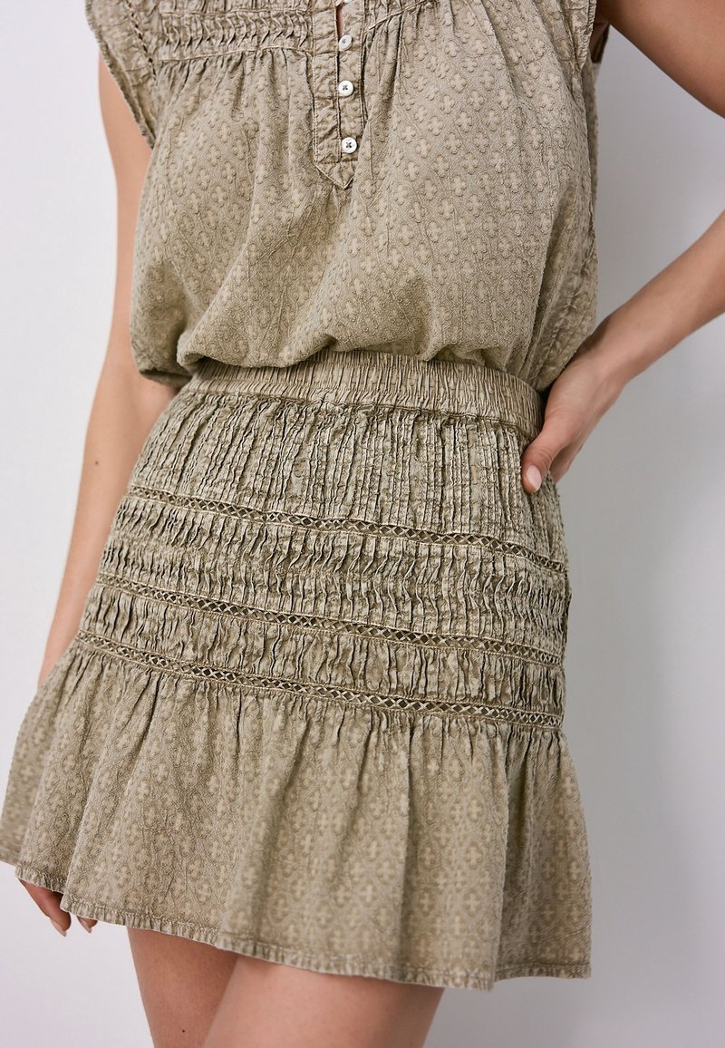 Femme portant une robe sans manches beige texturée avec taille smockée, inserts en dentelle et ourlet à volants, debout avec une main sur la hanche.