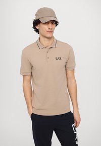 Beige polo med svarte detaljer og EA7-logo, sammen med en matchende caps. Modellen står med hendene i lomma, iført mørke bukser.