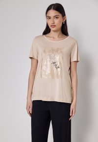 Joven mujer vestida con una camiseta beige de manga corta con un estampado cuadrado brillante y el texto negro "Toma el Riesgo", combinada con pantalones negros, se encuentra de pie contra un fondo liso.