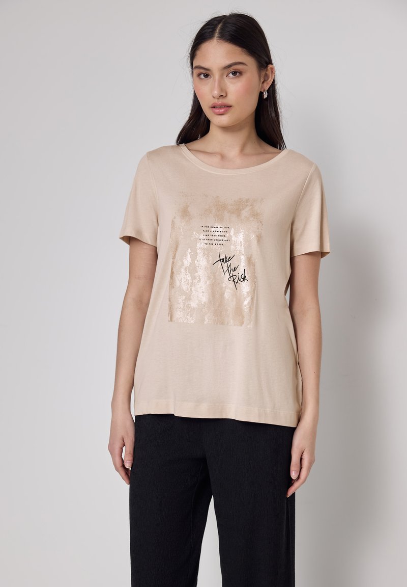 Joven mujer vestida con una camiseta beige de manga corta con un estampado cuadrado brillante y el texto negro "Toma el Riesgo", combinada con pantalones negros, se encuentra de pie contra un fondo liso.