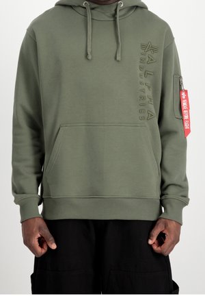Bluza z kapturem Alpha Industries w oliwkowym kolorze, z kieszenią kangurką z przodu, sznurkami, kieszenią na zamek na rękawie oraz czerwoną metką „Remove Before Flight” na rękawie.