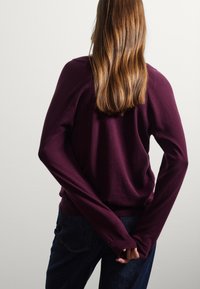 Bordeauxfarbener Pullover mit langen Ärmeln, lässiger Passform und geripptem Saum, kombiniert mit dunkelblauen Denim-Jeans, von hinten betrachtet.