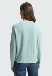 Hellblaue, strukturierte Bluse mit langen Ärmeln, lockerem Schnitt und weitem Halsausschnitt; kombiniert mit dunklen Jeans, von hinten angezeigt.
