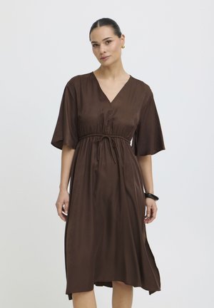 Naine, kes kannab pruuni V-kaelusega midi-kleit lühikeste laiade varrukatega ja paelaga vöökohal, seisab tavalise hele tausta ees.