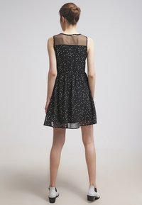 Robe noire sans manches avec des pois blancs, panneau supérieur transparent, taille élastique et jupe évasée. L'ourlet tombe au milieu des cuisses.
