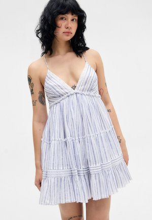 Młoda kobieta o czarnych kręconych włosach, ubrana w białą i niebieską sukienkę w paski bez rękawów z cienkimi ramiączkami, pokazująca wiele tatuaży na ramionach.