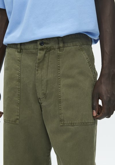 Pantalon en coton vert olive avec une fermeture à boutons, doté de poches latérales et d'une coupe droite. Porté avec un t-shirt bleu clair.