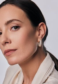 Złote kolczyki w kształcie obręczy z plecionym wzorem i małymi kryształowymi akcentami. Modelka ma średnio brązową cerę, delikatny makijaż i proste ciemne włosy.