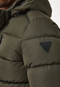 Olivgrüne Pufferjacke aus strukturiertem Material mit horizontalen Nähten, ausgestattet mit einer Reißverschlusstasche und einem schwarzen Logo-Patch auf der Schulter.