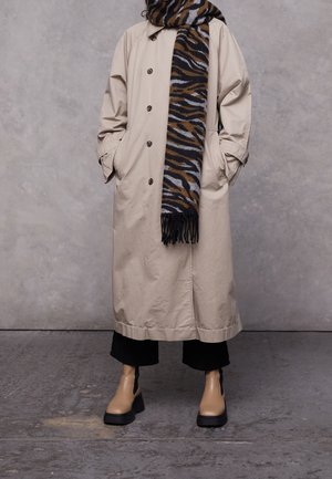 Personne portant un trench-coat beige, un pantalon noir court, des bottines marron clair et une écharpe imprimée animal en marron, noir et gris.