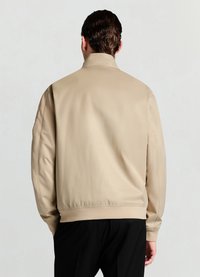 Veste bomber beige à texture lisse, col montant, poignets et ourlet élastiques, avec une coupe décontractée et un design minimaliste.