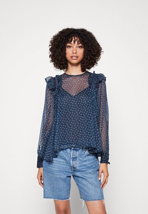 Blusa in rete blu navy con motivi floreali, colletto e maniche volant, abbinata a pantaloni corti di jeans. Texture trasparente e vestibilità comoda.