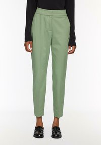 Groene tailleurbroek met een hoge taille, taps toelopende broekspijpen en zijzakken, gemaakt van een gladde stof. Draagt met zwarte schoenen.