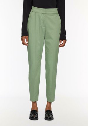 Pantalons ajustés verts avec une taille haute, des jambes fuselées et des poches latérales, fabriqués dans un tissu lisse. Portés avec des chaussures noires.