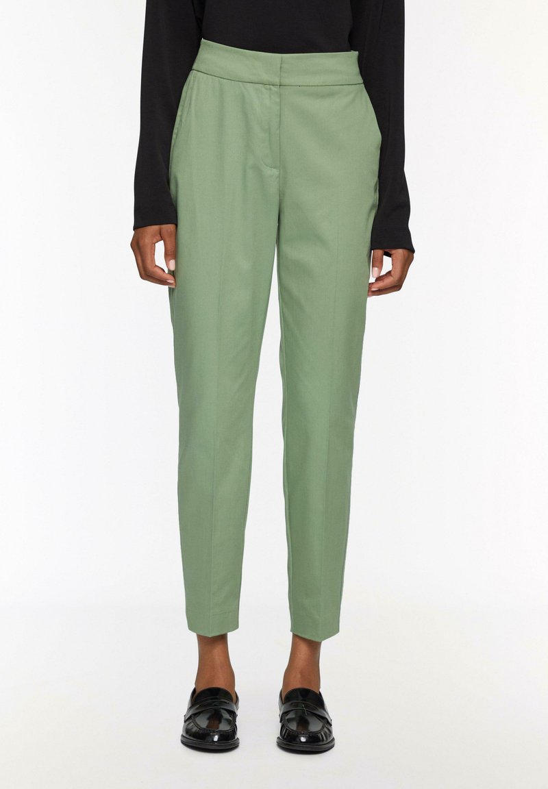 Groene tailleurbroek met een hoge taille, taps toelopende broekspijpen en zijzakken, gemaakt van een gladde stof. Draagt met zwarte schoenen.