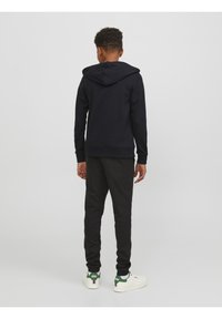 Zwarte hoodie met een grote capuchon, gecombineerd met donkergrijze joggers. De outfit heeft ribgebreide manchetten en wordt aangevuld met witte sneakers met groene accenten.