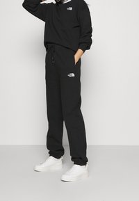 Schwarzer Kapuzenpullover und Jogginghose aus weichem Baumwollstoff. Mit einem Logo auf der Brust und der Tasche der Hose. Getragen mit weißen Sneakers.