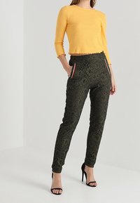 Żółty prążkowany top z długim rękawem w połączeniu z czarnymi spodniami o taperowanym kroju z wzorem koronkowym, posiadającymi boczne kieszenie oraz cienkie czerwone akcenty. Czarne sandały na wysokim obcasie.