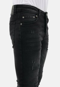 Zwarte versleten jeans met een slim fit, zichtbare scheuren in de stof en zilveren hardware bij de voorkeurs en de tailleband.
