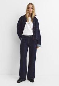 Giacca di jeans scura con grandi tasche e bottoni argentati, abbinata a una maglietta bianca e jeans larghi scuri. Scarpe nere con punta arrotondata.
