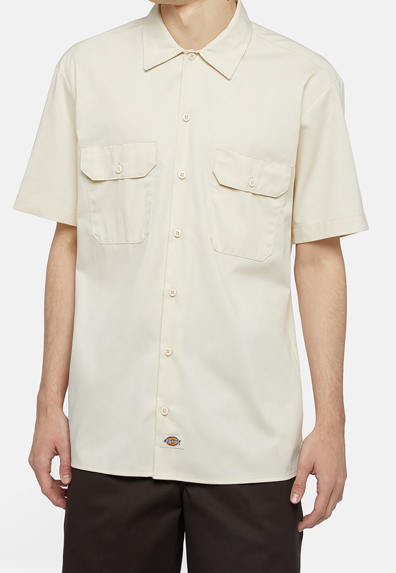 Camicia a maniche corte di colore beige chiaro realizzata in tessuto morbido, con due tasche sul petto, sei bottoni e un piccolo logo sull'orlo inferiore.