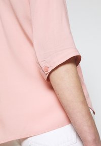 Short-sleeve, blush pink shirt with a textured pattern, featuring a button detail on the cuff and a straight hem, paired with white pants.