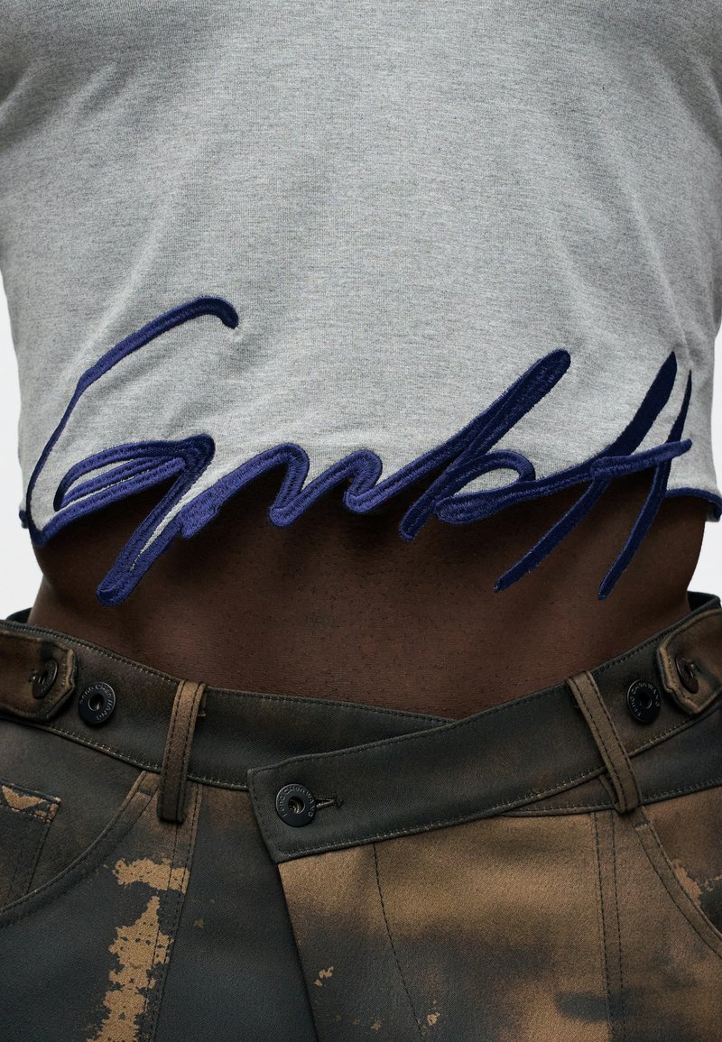 Grijze katoenen t-shirt met blauwe geborduurde tekst aan de onderrand, gecombineerd met donkere, gestructureerde broeken met een vervaagd patroon en knoopsluiting.