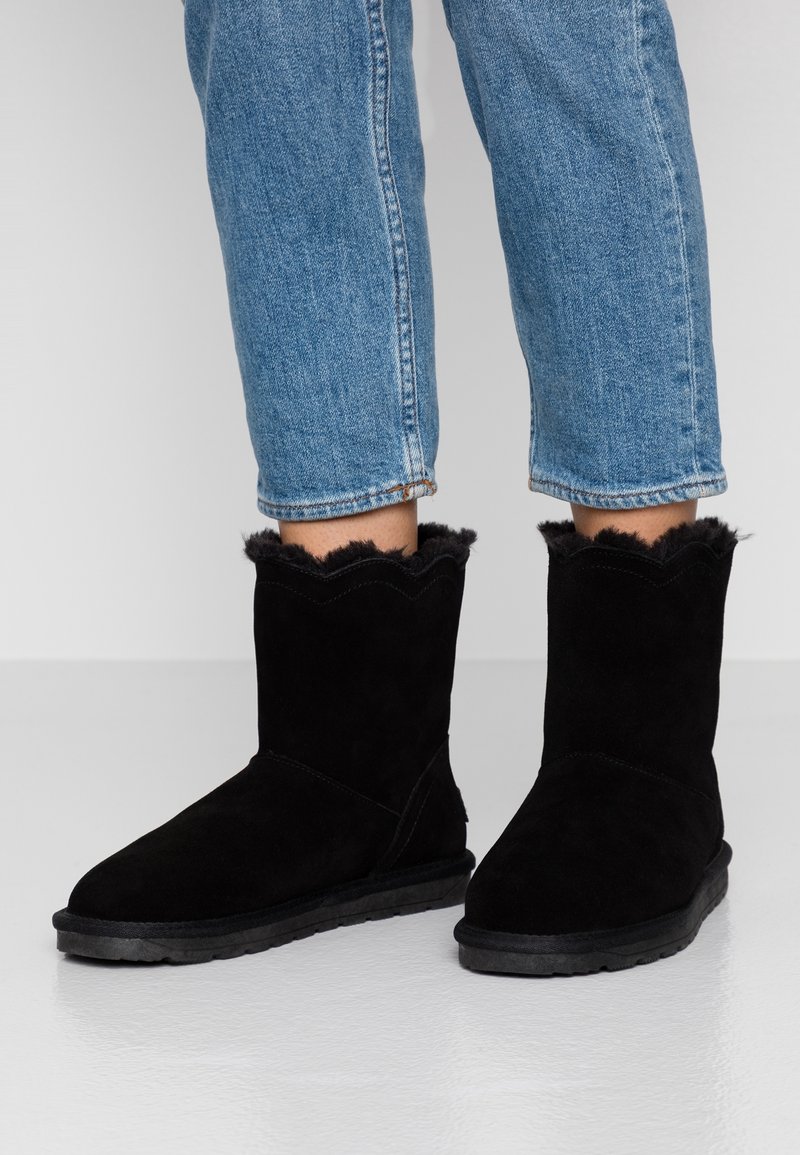 Bottes en daim noir arrivant à la cheville avec doublure en fourrure portées avec un jean bleu court sur un sol clair et lisse.