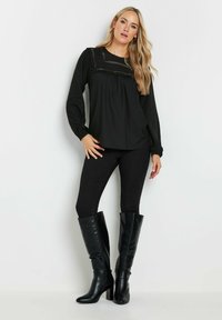 Blusa nera a maniche lunghe con dettagli a volant e cuciture decorative, abbinata a jeans skinny neri e stivali alti in pelle nera.