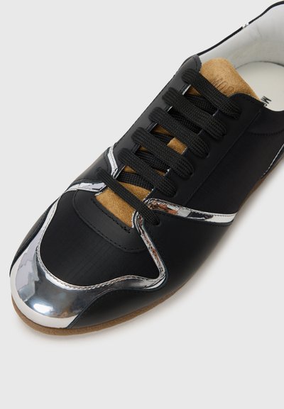 Black sneaker with a shiny silver toe cap, textile upper, and tan suede accents. Features black laces and a cushioned collar for comfort.
