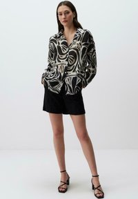 Blusa a maniche lunghe in tessuto trasparente con un motivo astratto in bianco e nero. Abbinata a pantaloni corti neri e sandali con cinturini neri.