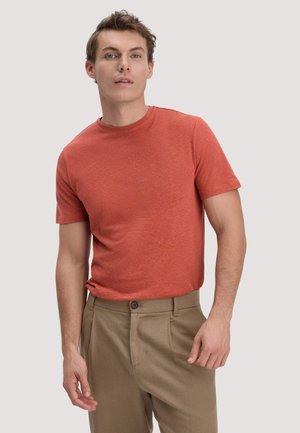Mann trägt ein enganliegendes, kurzärmeliges T-Shirt in Rostfarbe und beige Hose, steht vor einem schlichten hellen Hintergrund.
