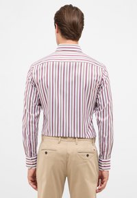 Camicia a maniche lunghe da uomo con righe verticali in rosso scuro e bianco. Presenta un colletto classico e polsini con bottoni; indossata con pantaloni khaki.