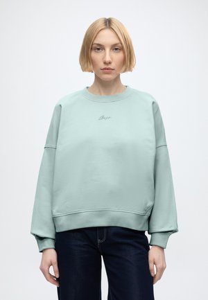 Młoda kobieta z krótkimi blond włosami, ubrana w jasnoniebieską, oversize bluzę i ciemne dżinsy, stojąca na jednolitym białym tle.