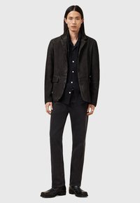 Blazer de cuero negro con un diseño estructurado, que presenta un cierre de botón único, tres bolsillos frontales y un cuello de muesca, combinado con pantalones oscuros.