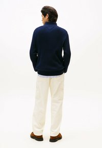 Navyblaue, gerippte Strickjacke mit hohem Kragen, getragen über einem weiß-blau gestreiften Hemd, kombiniert mit beigen Cargo-Shorts und braunen Schuhen.