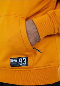 Hand in der vorderen Tasche eines orangen Sweatshirts mit einem "PROTEST 93"-Patch und passendem Reißverschlussanhänger sichtbar.