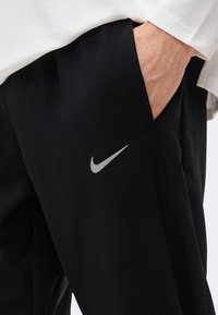 Χέρι στην τσέπη μαύρου παντελονιού Nike με ανακλαστικό λογότυπο swoosh, συνδυασμένο με λευκό μακρυμάνικο πουκάμισο που φαίνεται στη μανσέτα.