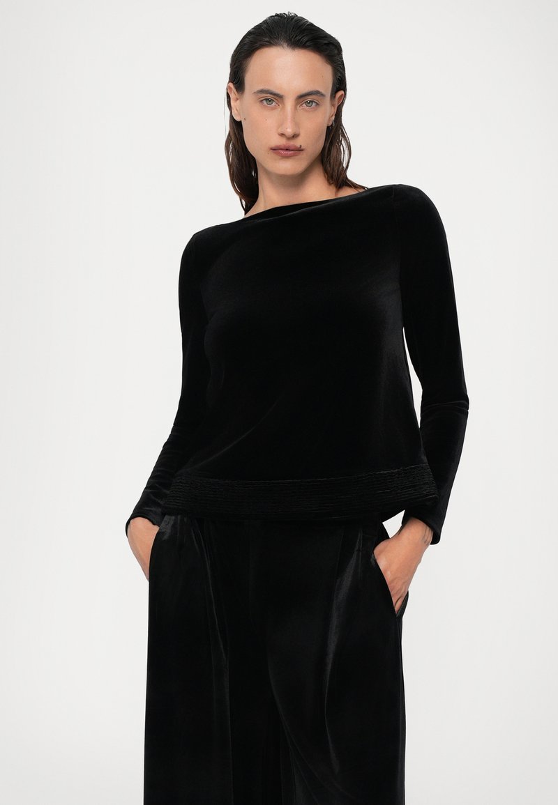 Black velvet long-sleeve top with a wide neckline and textured waistband. Model poses with hands in pockets, showcasing soft fabric and relaxed fit.
