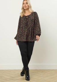 Schwarze Bluse mit Blumenmuster, rundem Ausschnitt und Bindedetail, langen Ärmeln und Peplum-Saum, kombiniert mit schwarzen Hosen und glänzenden Ankle Boots.