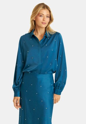 Conjunto de blusa y falda en color azul verdoso, confeccionado en una tela suave, con adornos similares a perlas esparcidos. Manga larga con puños abotonados.