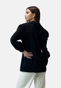 Camisa negra de gran tamaño hecha de tejido texturizado, con cuello y mangas largas y holgadas. Combinada con pantalones blancos.