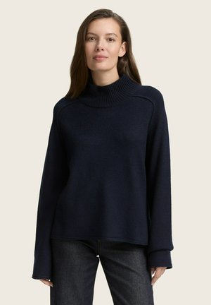 Pull-over col roulé en tricot navy avec des manches longues et une texture côtelée ; présente une coupe décontractée et un large col roulé ; porté avec un jean foncé.
