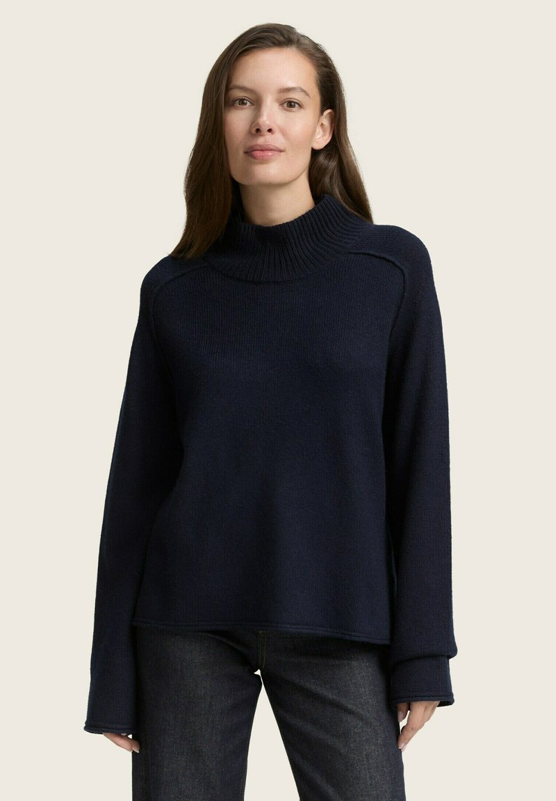 Maglione a collo alto in maglia blu navale con maniche lunghe e texture a coste; presenta una vestibilità rilassata e un ampio colletto arrotolato; indossato con jeans scuri.