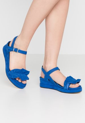 Blue suede sandals with a wide ruffled strap, ankle buckle, and a wedge heel design. The sole is textured for grip.